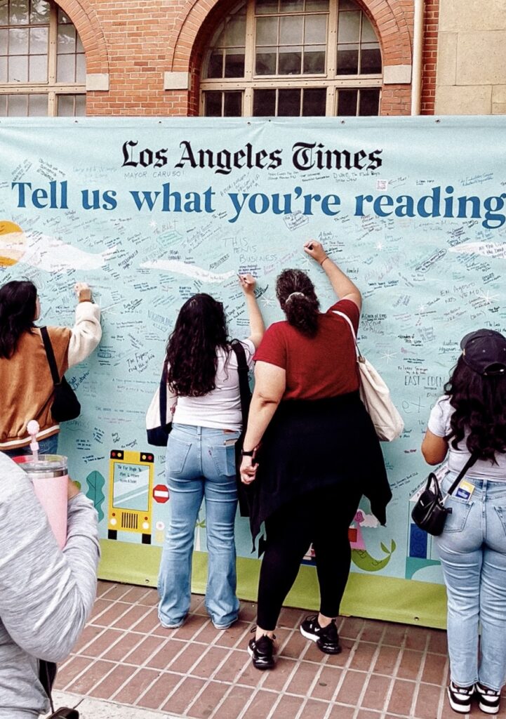 Festival of book visitors, outside, signing a large poster, pinned against a wall that says, "Los Angeles Times, Tell us what your reading".