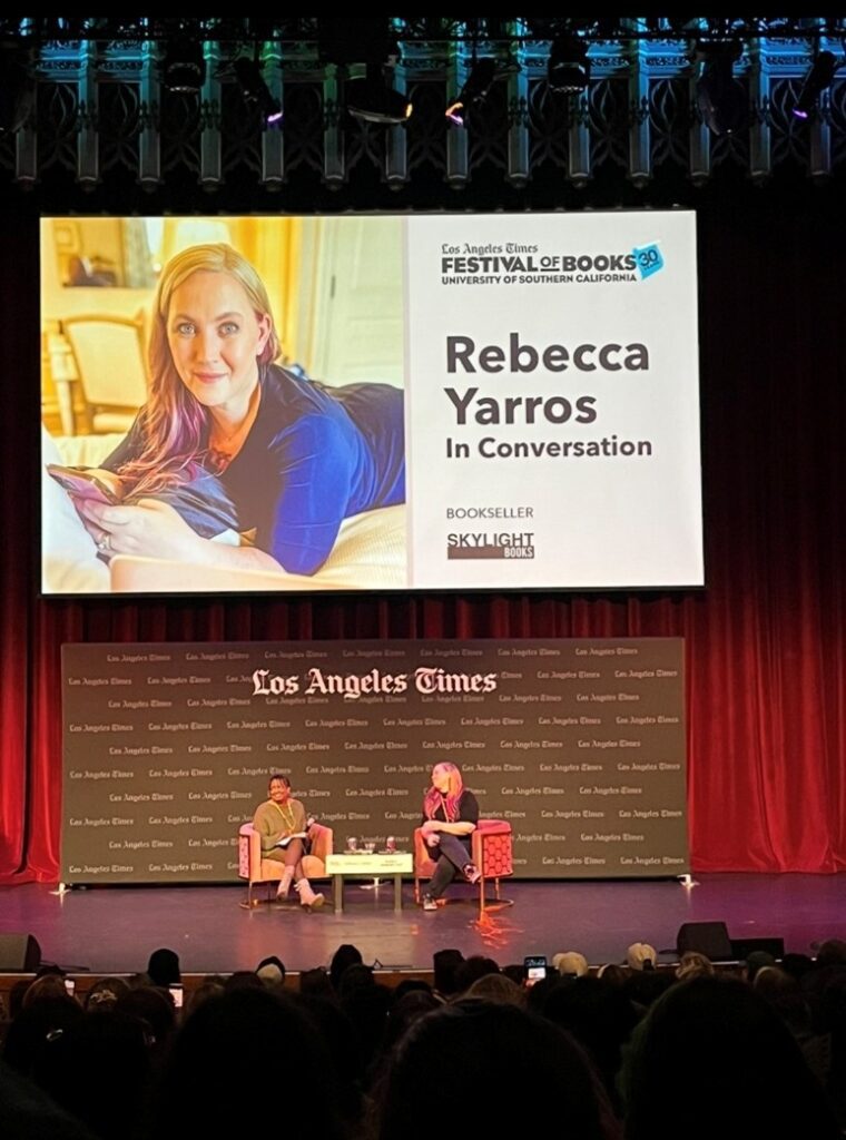 Image of Rebecca Yarros sitting on a stage with a large screen in the background. The image is split into two sections. The left section is showing an image of her laying on a bed in a brightly lit room with her phone in her hand. She's wearing a blue shirt, has long blond hair with pink tips and is smiling at the camera. On right side there is text that reads: "Los Angeles Times Festival of Books University of Southern California, Rebecca Yarros, In Conversation, Bookseller, Skylight Books.